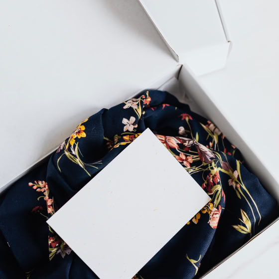 From above of stylish black silk floral pattern cloth with white visit card mockup placed in white carton box after receiving postal delivery of online order